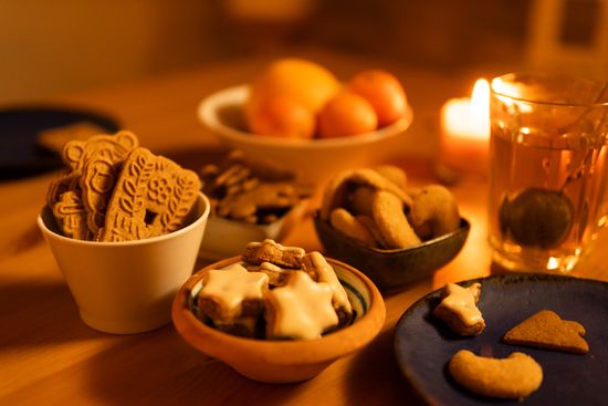 KI generiert: Das Bild zeigt eine gemütliche Szene mit verschiedenen Weihnachtsplätzchen in Schalen und einem Glas Tee bei Kerzenlicht. Im Hintergrund sind Schalen mit Früchten zu sehen.