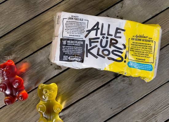 KI generiert: Das Bild zeigt eine Packung Toilettenpapier mit dem Aufdruck "ALLE FÜR KLOS!" auf einem Holzboden. Daneben stehen zwei große, bunte Gummibären.