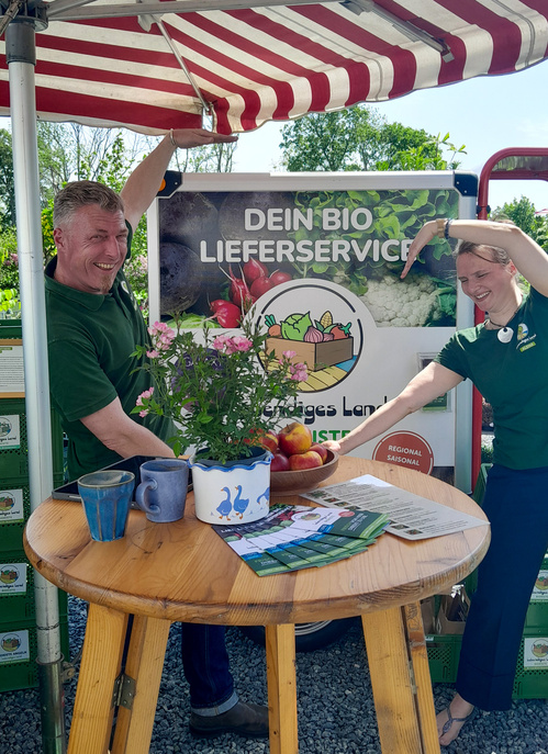 Christian und Alena freuen sich an einem Stand mit dem Schild "Dein Bio Lieferservice". Flyer und Blumen auf dem Tisch.