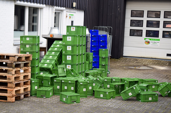 KI generiert: Grüne Kisten und Paletten stapeln sich vor einem Gebäude. Text: "Ansi Biohofladen" und "Lieferung - Biologische Kisten."