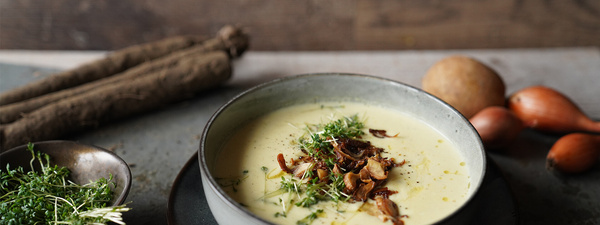 KI generiert: Das Bild zeigt eine Schüssel mit einer cremigen Suppe, garniert mit Kräutern und einigen Pilzen. Im Hintergrund liegen weitere Zutaten wie schwarzwurzelähnliches Gemüse, Kartoffeln und Schalotten.