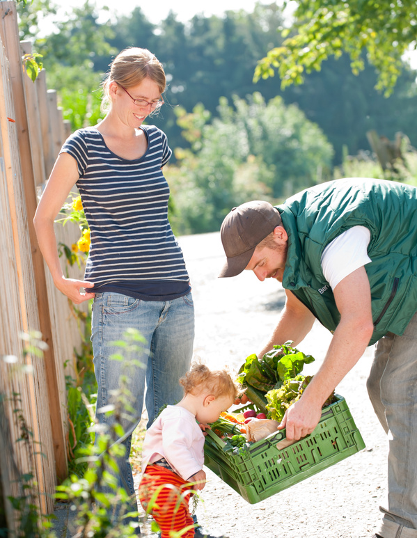 KI generiert: Ein Mann überreicht einer Frau eine Gemüsekiste im Freien. Ein Kind steht daneben.