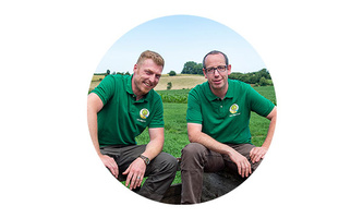 KI generiert: Zwei Männer in grünen Poloshirts sitzen lächelnd auf einer Wiese. Im Hintergrund ist eine Landschaft zu sehen.