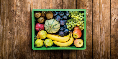 KI generiert: Kiste mit verschiedenen Obstsorten: Bananen, Äpfel, Melone, Trauben, Kiwi und Pflaumen auf Holzhintergrund.