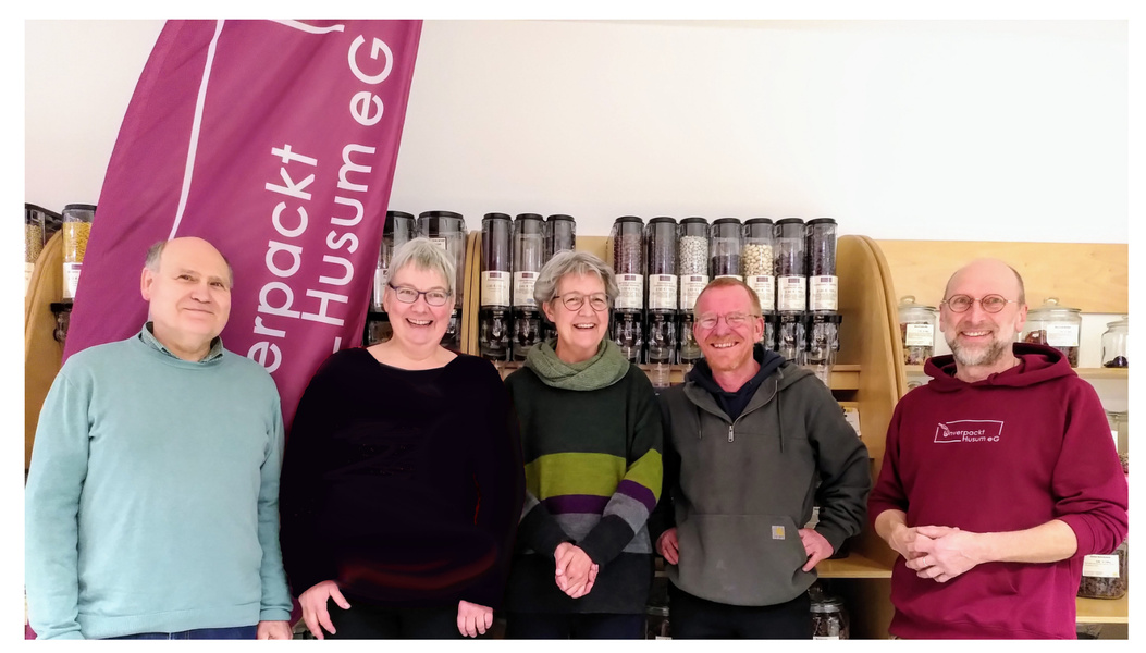 KI generiert: Eine Gruppe von fünf Personen steht vor Regalen mit Behältern. Text: "unverpackt Husum eG".