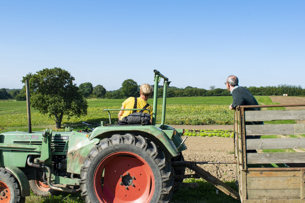 KI generiert: Zwei Personen auf einem Traktor-Anhänger blicken über ein weites Feld.