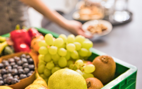 KI generiert: Eine grüne Kiste mit frischem Obst wie Trauben, Birnen und Kiwis. Im Hintergrund eine Hand mit einer Schale.