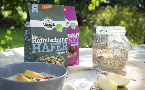 KI generiert: Müslipackungen von Bauckhof und eine Schale mit Müsli und Apfelstücken auf einem Tisch im Freien.