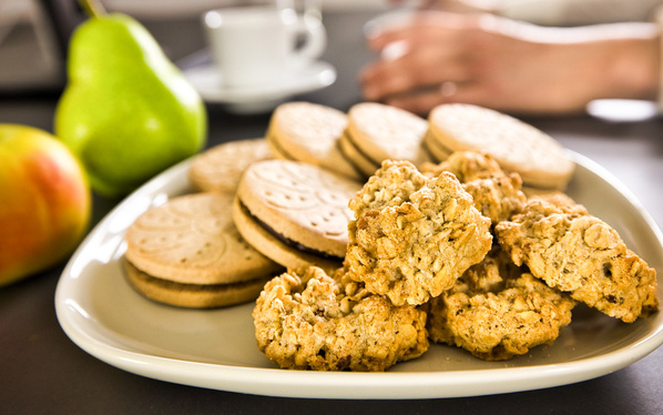 KI generiert: Eine Platte mit Keksen und Obst, darunter ein Apfel und eine Birne. Im Hintergrund Hände und eine Tasse.