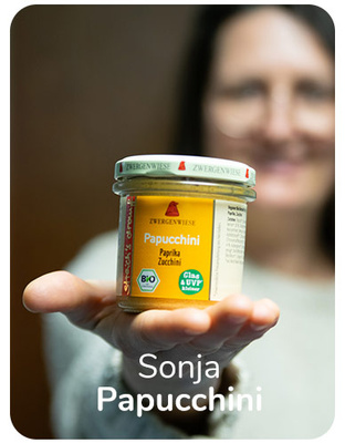 KI generiert: Eine Frau hält ein Glas "Papucchini Paprika Zucchini" von Zwergenwiese in die Kamera. Text: "Sonja Papucchini".