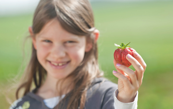 KI generiert: Mädchen hält eine Erdbeere lächelnd in der Hand; unscharfer grüner Hintergrund.
