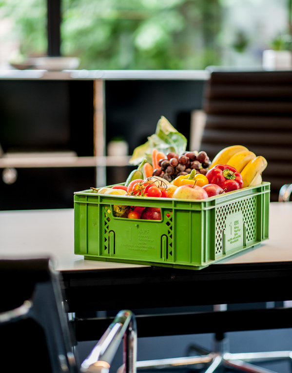 KI generiert: Grüne Kiste mit buntem Obst und Gemüse auf einem Tisch in einem Büro.