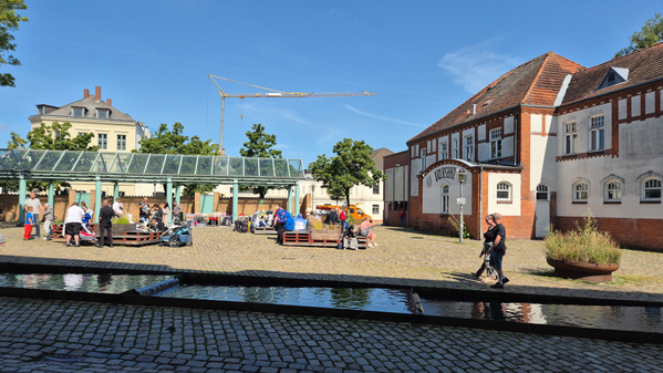 KI generiert: Menschen auf einem sonnigen Platz mit Cafés und einem Gebäude im Hintergrund. Text: "HAFEN SULZBACH".