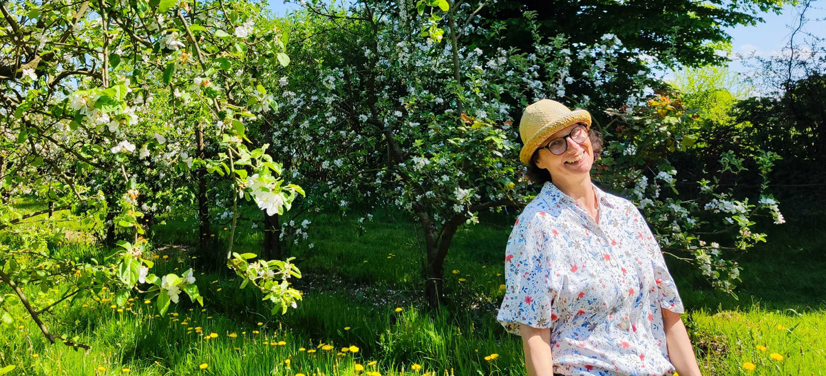 KI generiert: Eine Person mit Sonnenhut steht lächelnd in einem blühenden Obstgarten.