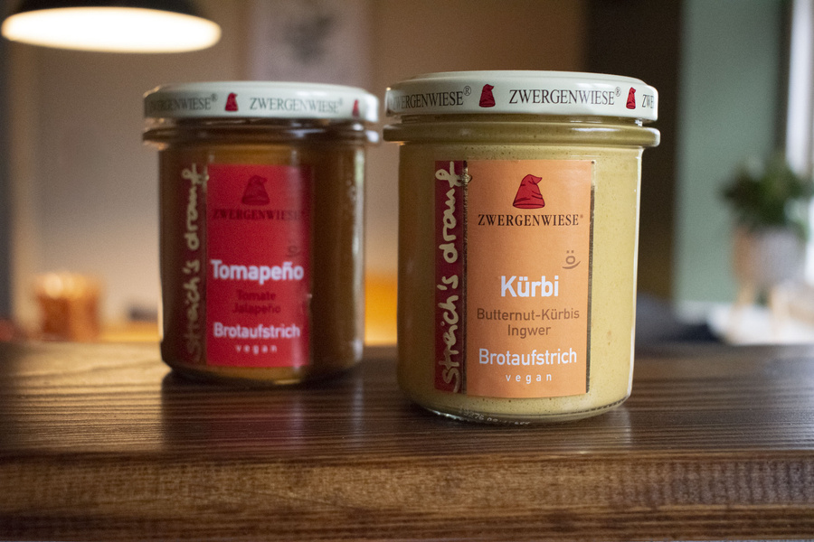 KI generiert: Zwei Zwergenwiese Brotaufstriche im Glas auf einem Tisch. Texteinblendung: „Tomapeño“ und „Kürbi“.