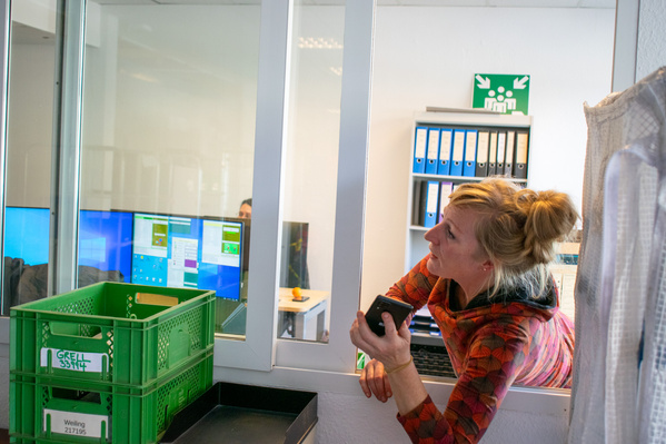 KI generiert: Eine Frau lehnt entspannt an einer Trennwand, hält ein Smartphone. Im Büro sind grüne Kisten und Büroordner.