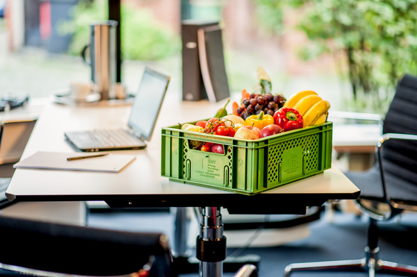 KI generiert: Eine grüne Kiste voller Obst steht auf einem Bürotisch mit Laptop, Ordner und Stuhl.