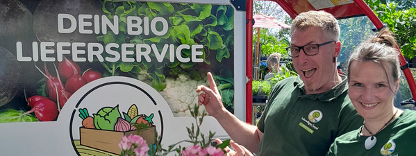 Christian und Alena stehen vor einem Schild mit der Aufschrift "DEIN BIO LIEFERSERVICE".
