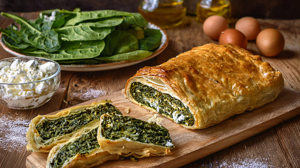 KI generiert: Blätterteigrolle mit Spinatfüllung, umgeben von frischem Spinat, Käse und Eiern auf einem Holztisch.