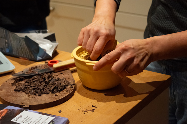 KI generiert: Hände zerkleinern Kekse in einer gelben Schüssel auf einem Tisch. Schokoladenspäne auf einem Brett daneben.