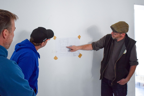 KI generiert: Drei Personen betrachten einen an der Wand befestigten Bauplan.