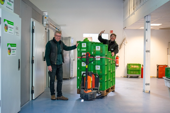 KI generiert: Zwei Personen posieren neben einem Stapel grüner Kisten auf einer Palette in einem Lagerraum.