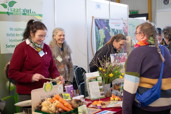 KI generiert: Menschen an einem Messestand für Saatgut und Pflanzenzucht. Text: "Wir züchten Vielfalt der Natur."