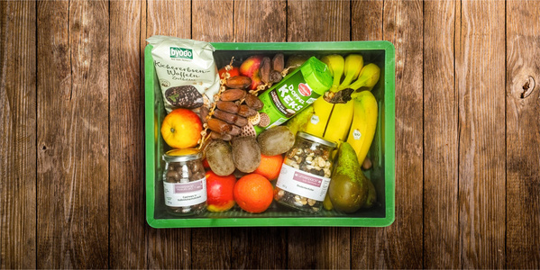 KI generiert: Eine Obst- und Gemüsekiste mit Bananen, Äpfeln, Kiwis, Nüssen und Snacks. Text: „Byodo Kichererbsen Waffeln“.