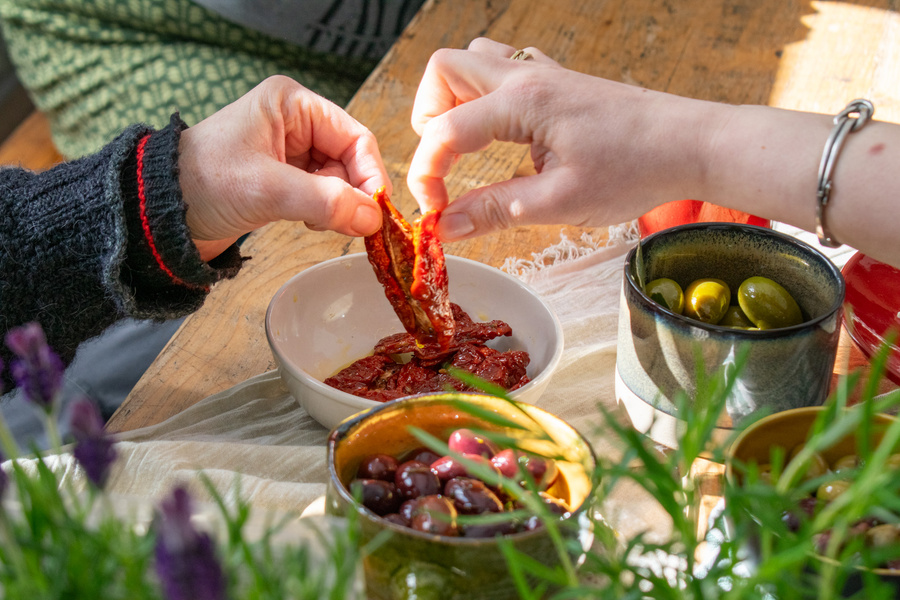 KI generiert: Hände nehmen getrocknete Tomaten aus einer Schüssel, umgeben von Oliven und anderen Snacks auf einem Holztisch.