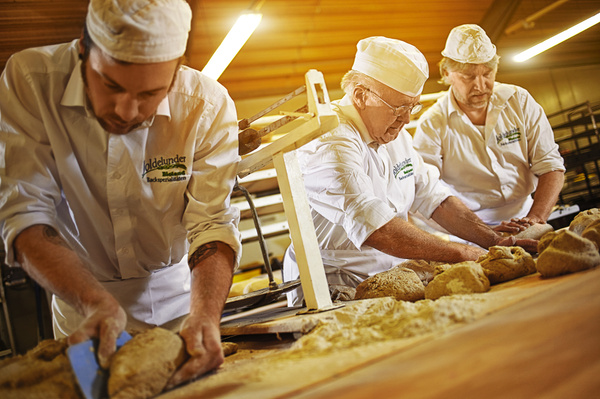 KI generiert: Bäcker formen Teig; traditionelle Handwerkskunst in einer Bäckerei. Text: Goldelunder Backparadies.