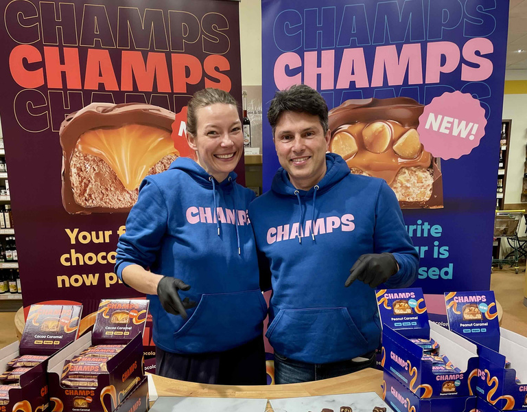 KI generiert: Zwei Personen stehen vor einem Tisch mit Schokoladenproben und tragen passende blaue Hoodies mit der Aufschrift "CHAMPS". Im Hintergrund befinden sich große Werbebanner für die Schokoladenmarke Champs.