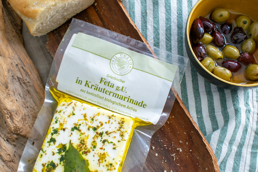 KI generiert: Verpackter Feta in Kräutermarinade neben Brot und einer Schale mit Oliven. Text: "Feta g.U. in Kräutermarinade".