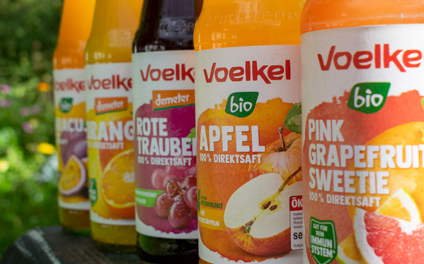 KI generiert: Fünf verschiedene Flaschen Voelkel-Säfte, darunter Apfel- und Grapefruitsaft, stehen aufgereiht im Freien.