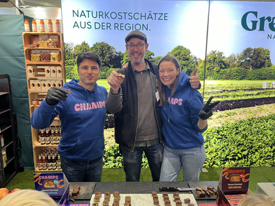 KI generiert: Das Bild zeigt drei Personen, die an einem Stand mit verschiedenen Snacks lächeln, möglicherweise auf einer Messe. Im Hintergrund befindet sich ein großes Plakat mit der Aufschrift "Naturkostschätze aus der Region."