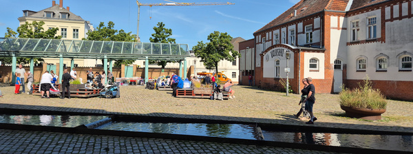 KI generiert: Menschen entspannen auf einem sonnigen Platz mit Pavillon und historischem Gebäude. Text: "VOLKSHAUS".