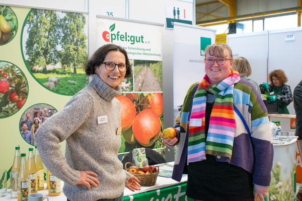 KI generiert: Zwei Personen vor einem Messestand von "apfel:gut e.V." mit Äpfeln und Infomaterial.