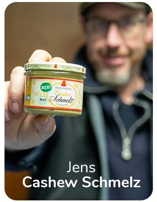 KI generiert: Ein Mann hält ein Glas mit der Aufschrift "Zwergenwiese Cashew Schmelz". Text im Bild: "Jens Cashew Schmelz".
