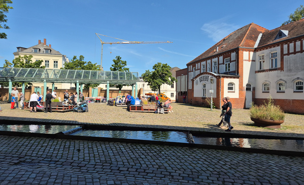 KI generiert: Ein belebter Platz mit Menschen auf Bänken, einem gläsernen Vordach und dem Gebäude "Schlachthof".