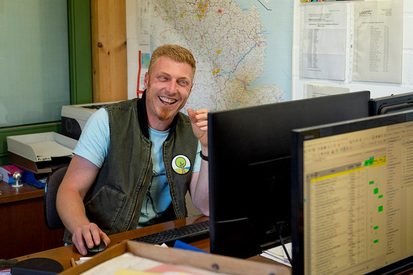 KI generiert: Ein Mann lächelt vor einem Computerbildschirm in einem Büro. Im Hintergrund hängt eine Landkarte.